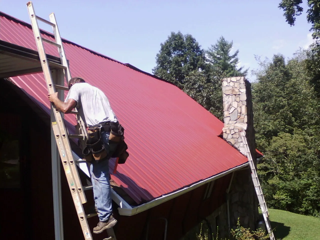 Roofer on a ladder beside a red metal roof and stone chimney — Soffit & Fascia Repair by Roofing Brothers in Brothers